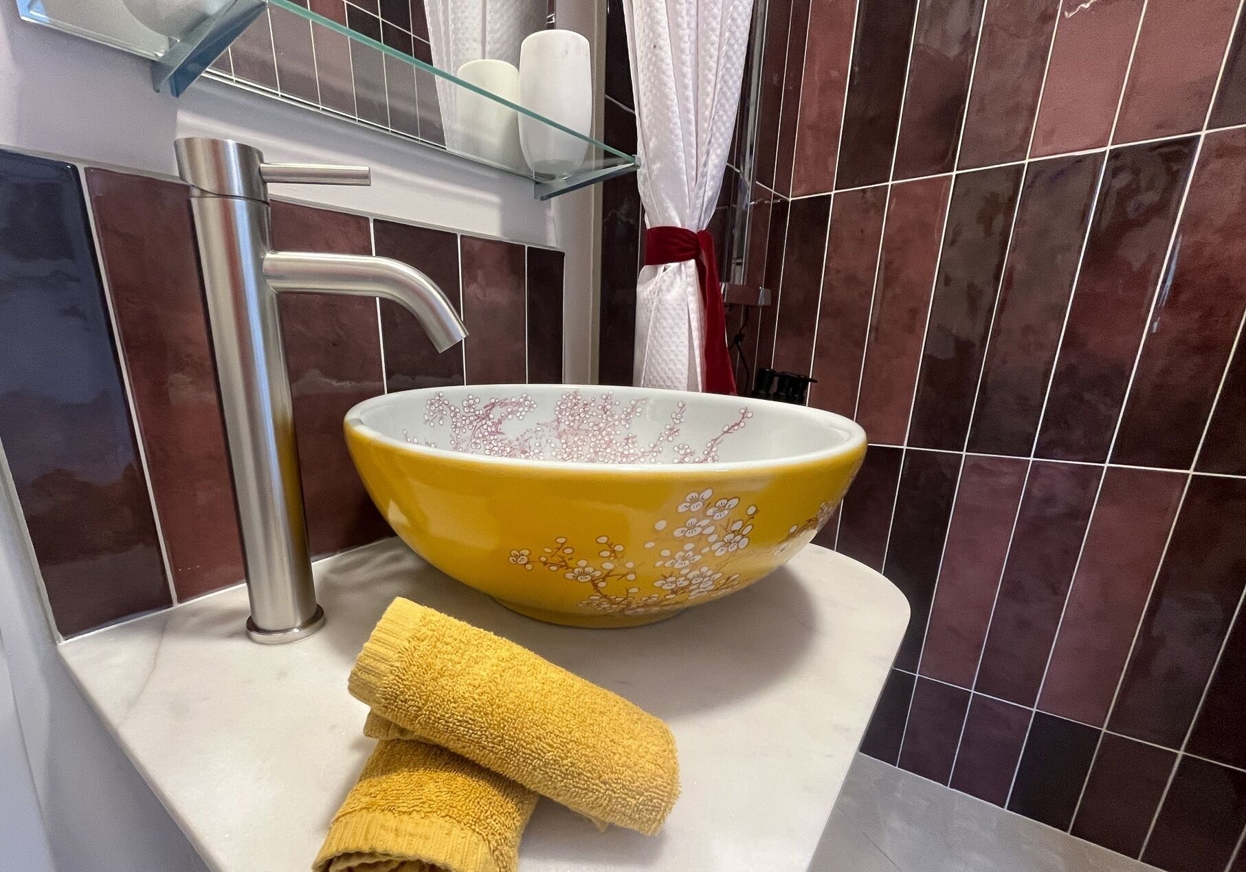 Une salle de bain avec un lavabo jaune, une étagère en verre où sont posés des articles de toilette et une serviette sur le comptoir.