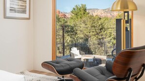 Ein modernes Hotelzimmer mit einem großen Fenster und Blick auf eine gebirgige Landschaft. Zwei schwarze Ledersessel und ein kleines Sofa sind vorhanden.