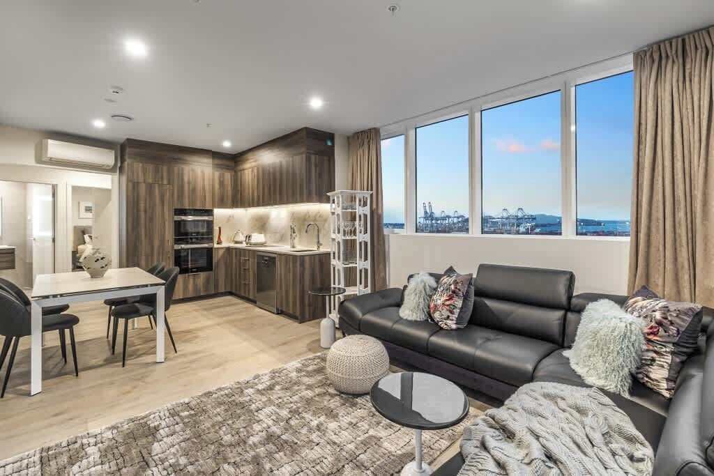 A modern living room with a black leather sofa, a dining table with chairs, and a kitchen area with wooden cabinets and a white island.