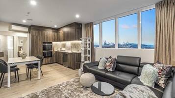 A modern living room with a black leather sofa, a dining table with chairs, and a kitchen area with wooden cabinets and a white island.