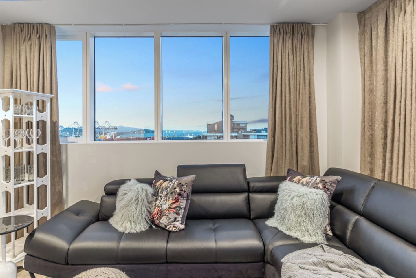 A modern living room with a black leather sofa, two decorative pillows, a white glass cabinet, and a large window offering a city view.