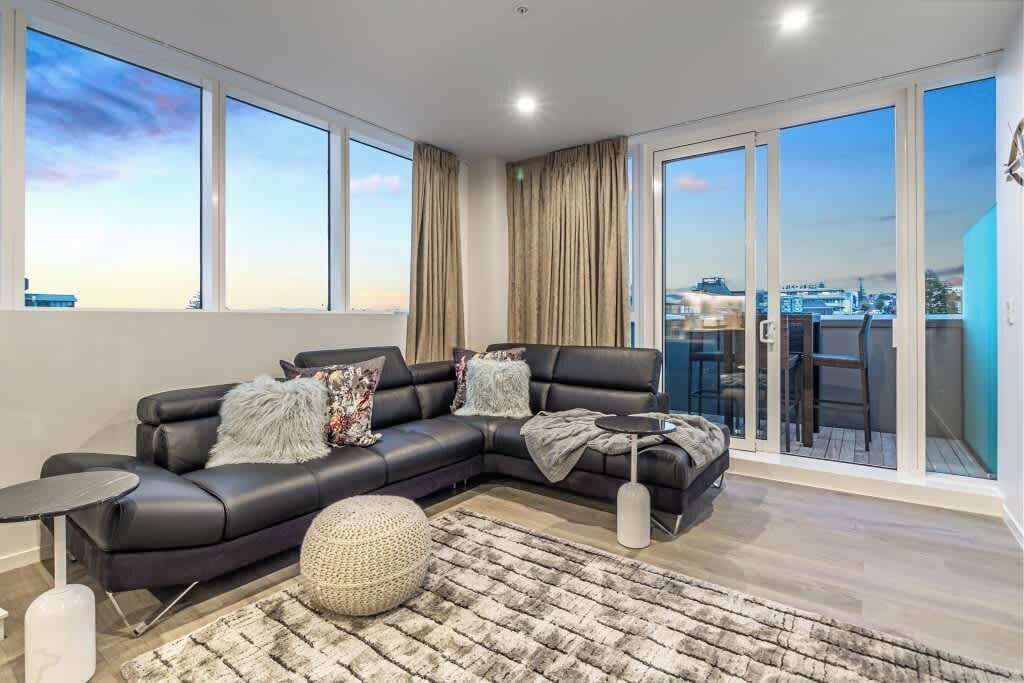 A modern living room with a black leather sofa, a round ottoman, and a small side table. Large windows offer a view of the cityscape.