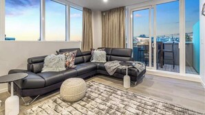 Una sala de estar moderna con un sofá de cuero negro, un taburete redondo y una pequeña mesita auxiliar. Amplias ventanas ofrecen vistas del paisaje urbano.