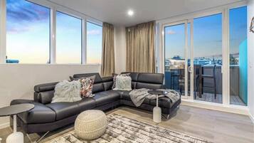 A modern living room with a black leather sofa, a round ottoman, and a small side table. Large windows offer a view of the cityscape.
