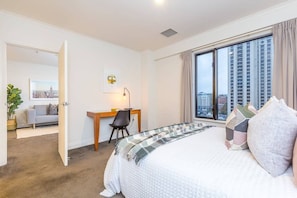 A hotel room with a bed, a desk, a chair, a window with a city view, and a door leading to another room. - Stylish Apt with Views Pool and Gym - Zodiak Stays (Auckland)