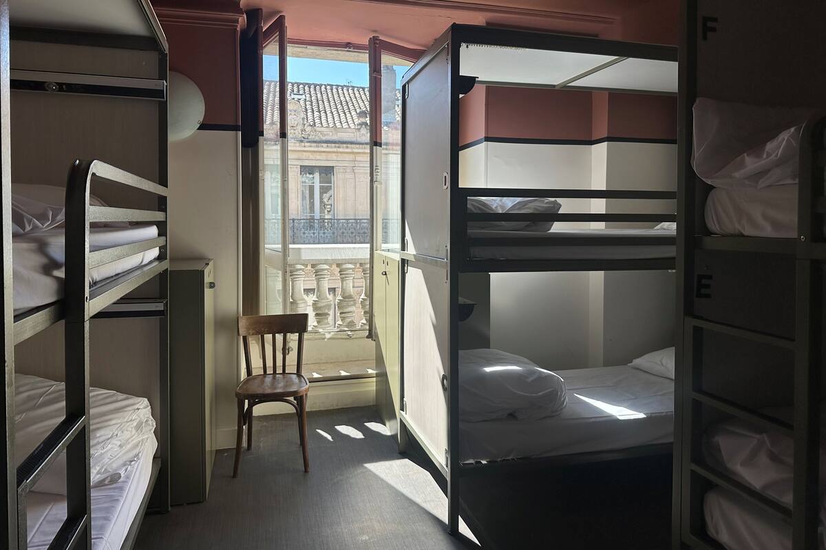 Een slaapzaal met stapelbedden, een stoel, en een balkon met uitzicht op gebouwen.