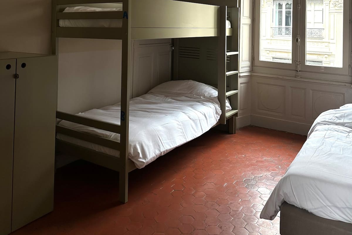 Een kamer met stapelbedden, een kledingkast, een decoratieve spiegel en een raam met uitzicht op gebouwen.