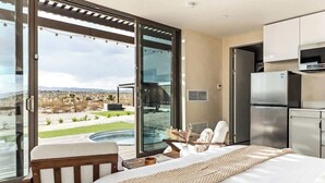 A modern bedroom with a large sliding glass door leading to a pool, a bed, a small table with chairs, and a kitchenette.