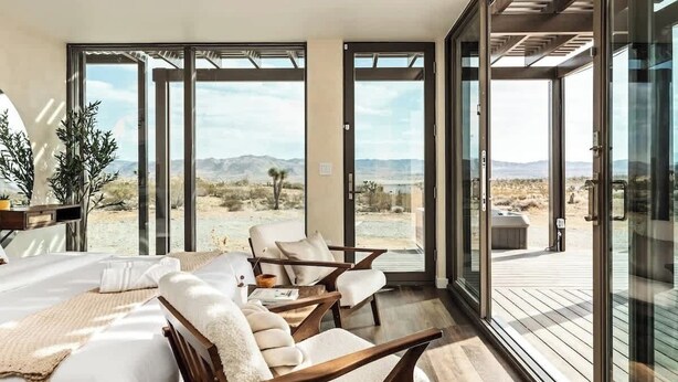 A modern room with a large bed, two chairs, and a view of a desert landscape through large glass doors.