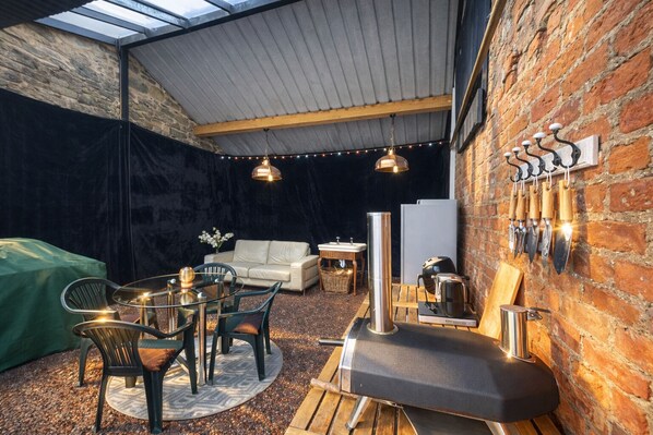 Un espace intérieur accueillant comprenant un coin repas, un canapé, un barbecue et un mur de briques orné d’outils suspendus.