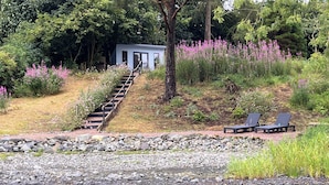 Une propriété au bord du lac, dotée d’une terrasse en bois, d’un chemin en pierre et d’un petit bâtiment avec une porte.