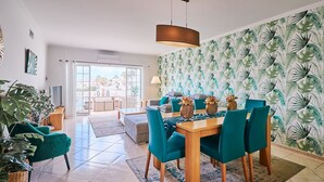 A modern living room with a dining area, featuring a large window, a ceiling fan, and a tropical wallpaper. - Apartment O Monte with pool and tennis court  (Guia)