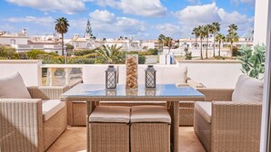A balcony with wicker furniture, a glass table, and a view of a coastal town with palm trees and white buildings. - Apartment O Monte with pool and tennis court  (Guia)