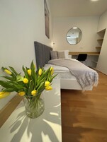 A modern bedroom with a bed, a bedside table with a vase of yellow tulips, a round mirror on the wall, and a wooden floor.