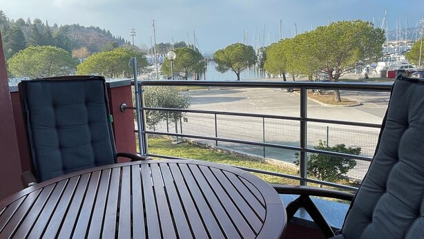 Een balkon met een ronde houten tafel en twee comfortabele stoelen, met uitzicht op een jachthaven met boten en een glooiend landschap.