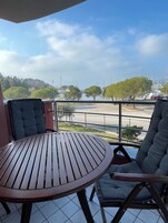 Un balcón con una mesa redonda de madera y dos sillas con cojines, con vistas a un puerto deportivo con barcos y un paisaje montañoso.