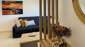 Een moderne woonkamer met een donkere bank, een bijzettafeltje, een vaas met gedroogde bloemen, een schilderij van een zonsondergang en een decoratieve spiegel.