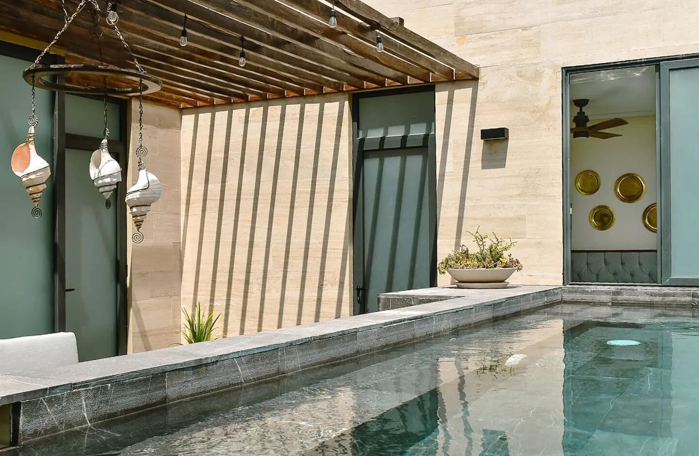 A modern outdoor pool area with a wooden pergola, a potted plant, and a glass door leading to a room with decorative plates.