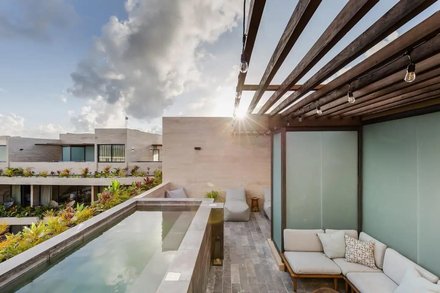 A modern outdoor pool area with a wooden pergola, a seating area with cushions, and a view of the surrounding buildings.