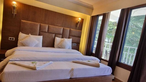 A double bed with a headboard, bedside lamps, and a view of greenery through large windows. - Hotel Taj palace Dalhousie (Dalhousie)