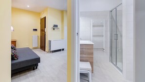 A modern hotel room with a bed, a small table, a radiator, a shower area, and a glass door leading to the bathroom. - Monteverde Cozy Apartment (Rome)