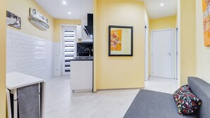 A modern kitchen with white cabinets, a black stove, and a white countertop. There is a dining area with a white table and a gray sofa. - Monteverde Cozy Apartment (Rome)