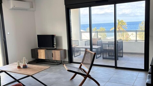 A modern living room with a ceiling fan, a TV, a dining area with a table and chairs, and a large sliding glass door leading to a balcony with a view of the ocean.
