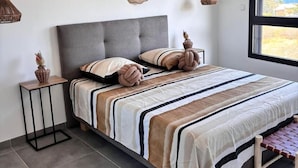 A modern bedroom with a grey upholstered bed, striped bedding, and a view of the outdoors through a large window. - Sunset - Front de mer (Trois-Bassins)