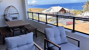 A balcony with two cushioned chairs and a table, overlooking a coastal view with buildings and palm trees. - Sunset - Front de mer (Trois-Bassins)
