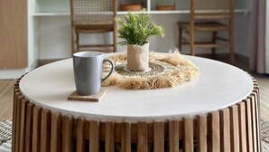 A round wooden table with a white top, a grey mug, and a small potted plant on a woven placemat. A round mirror hangs on the wall above a wooden desk with a woven chair. - Studio Eleven Alamanda Beach beachfront (Saint Martin)