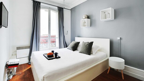 A neatly made bed with a tray of breakfast items, a bedside table, a small white stool, and a wall-mounted shelf with a decorative item. - Cosy 1br/4p - Montmartre/sacre Coeur (Paris)