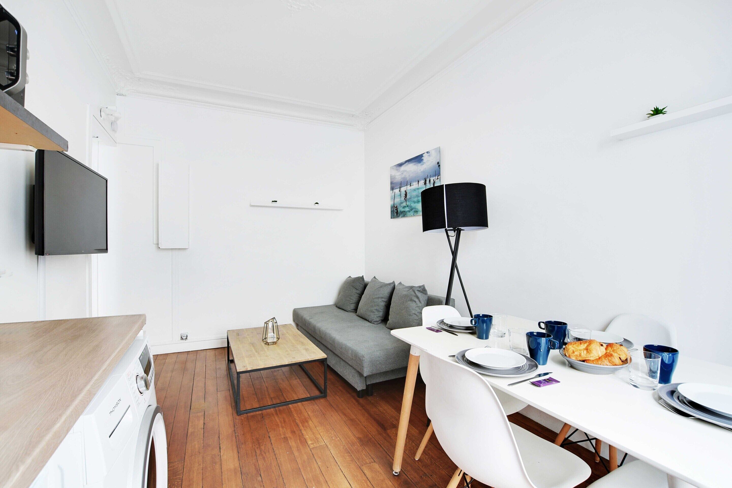 A compact living space with a washing machine, a grey sofa, a dining table with chairs, and a TV mounted on the wall.