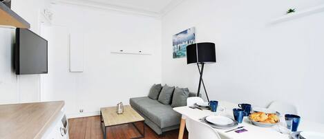 A compact living space with a washing machine, a grey sofa, a dining table with chairs, and a TV mounted on the wall.