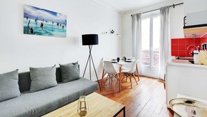 A modern living room with a grey sofa, a dining table with chairs, a floor lamp, and a kitchen area with red tiles. - Cosy 1br/4p - Montmartre/sacre Coeur (Paris)