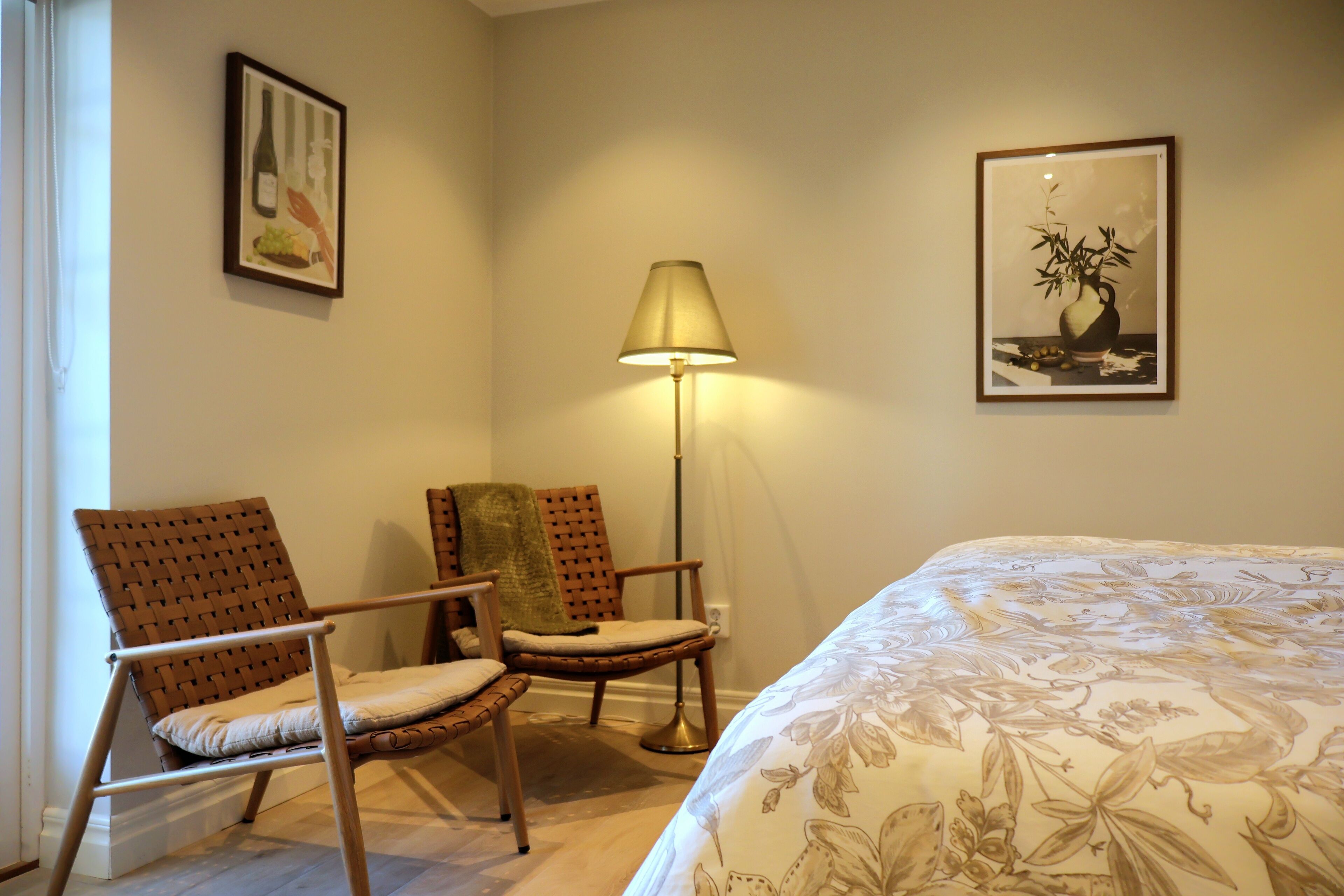 A bedroom with a bed, two chairs, a floor lamp, and two framed pictures on the wall.