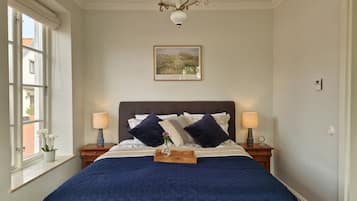 A bedroom with a large bed, two bedside lamps, a chandelier, a window with a view, and a framed picture on the wall.