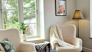 A room with two white armchairs, a floor lamp, a framed picture on the wall, and a window with a view of greenery outside. - Fontänens Bed & Breakfast (Trelleborg)
