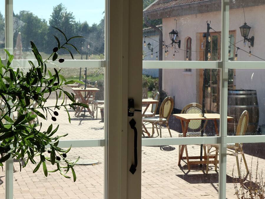 A view through a glass door of an outdoor patio area with tables and chairs, a potted plant, and a building with a tiled roof.