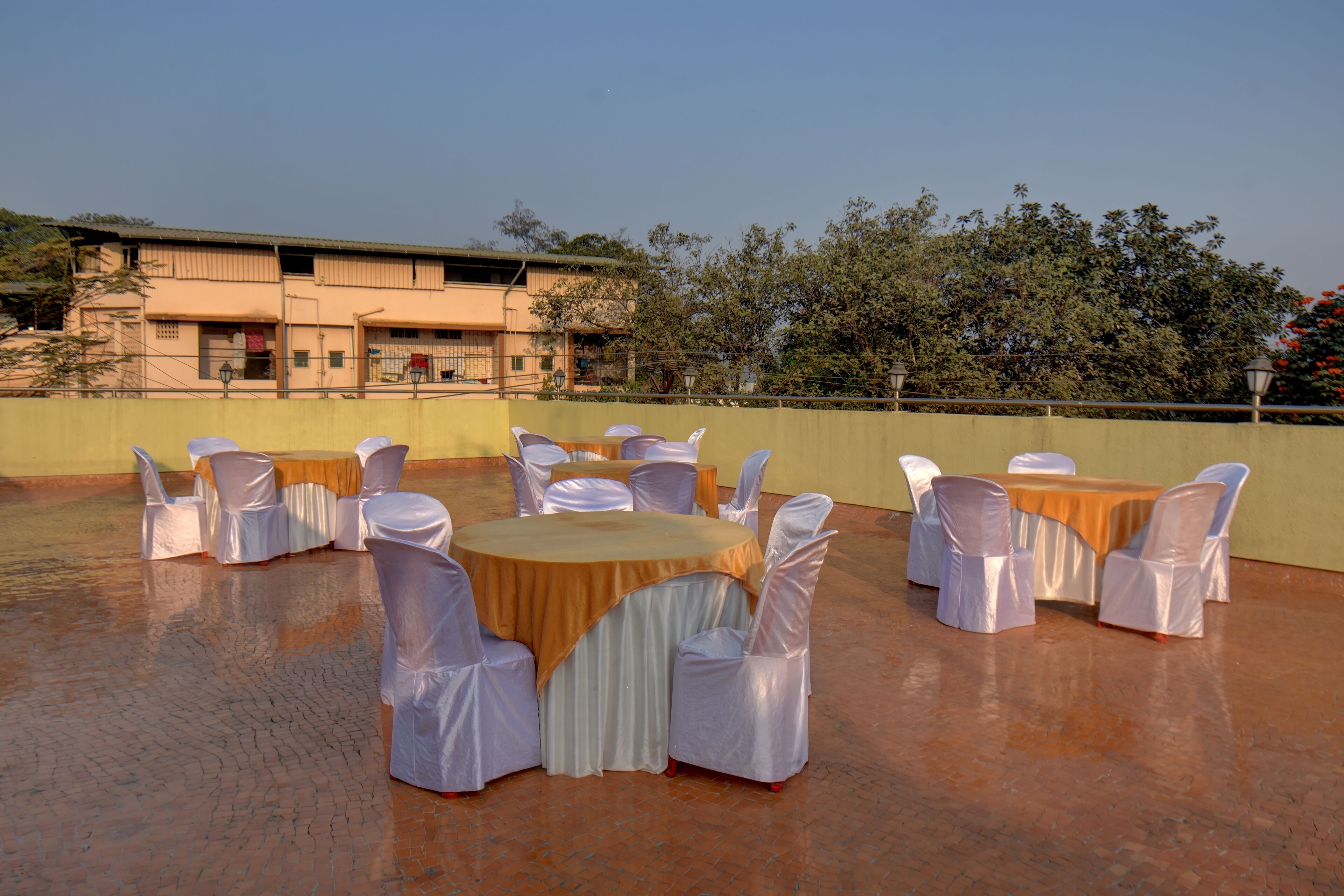 A rooftop terrace with round tables and chairs, a building in the background, and trees.