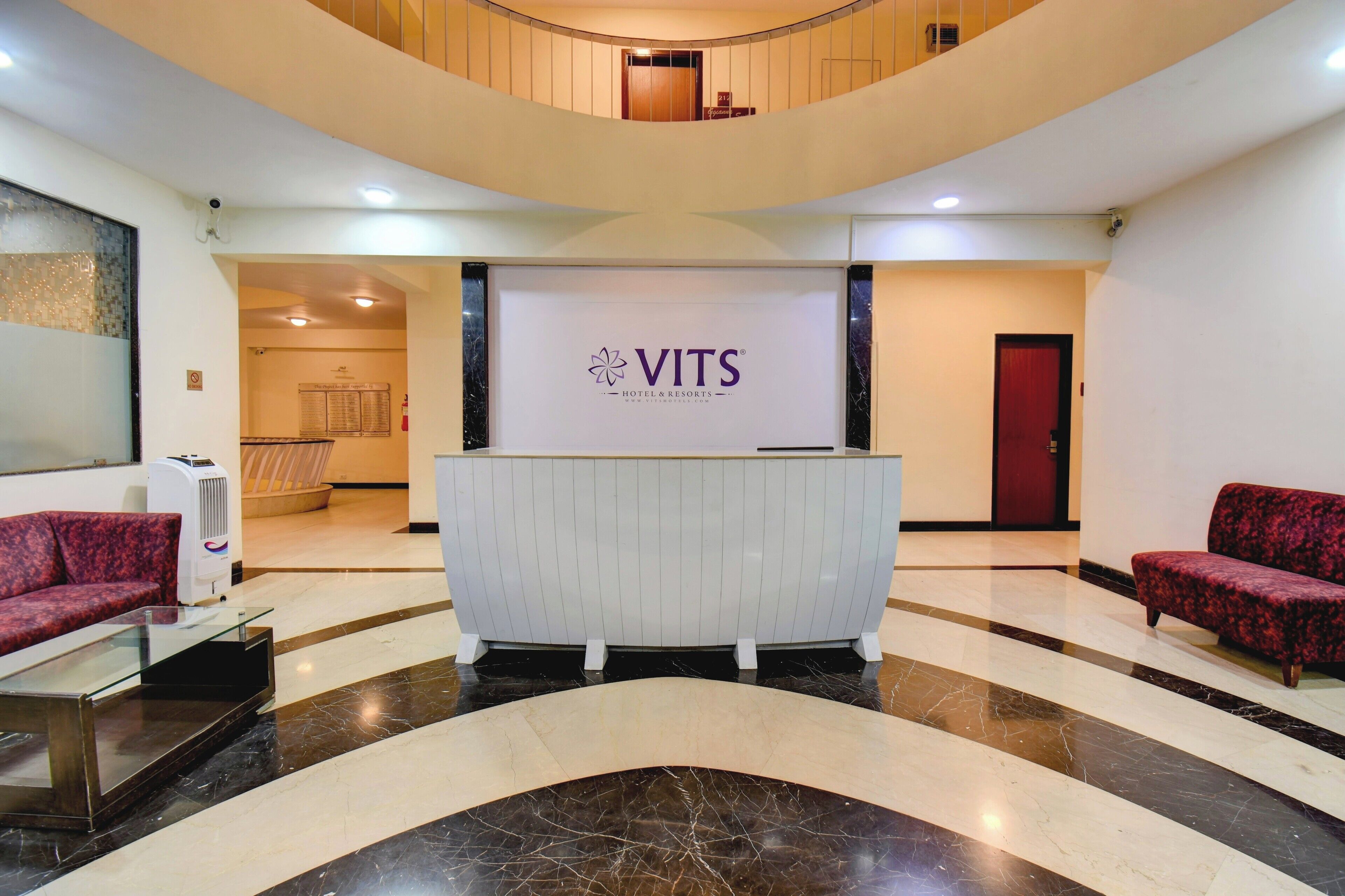 A reception area with a VITS Hotel & Resorts sign, a reception desk, a red sofa, and a glass coffee table.