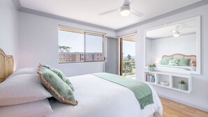 A bedroom with a large bed, a window with a view of buildings, a ceiling fan, and a wall-mounted shelf with books and plants. - Commodore 6, 6 Intrepid Cl - water views, Air Con, Wi-Fi (Nelson Bay)