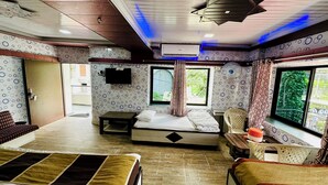 Ein Zimmer mit Bett, Fernseher, einem Fenster mit Vorhängen, einem Ventilator und Blick ins Grüne.