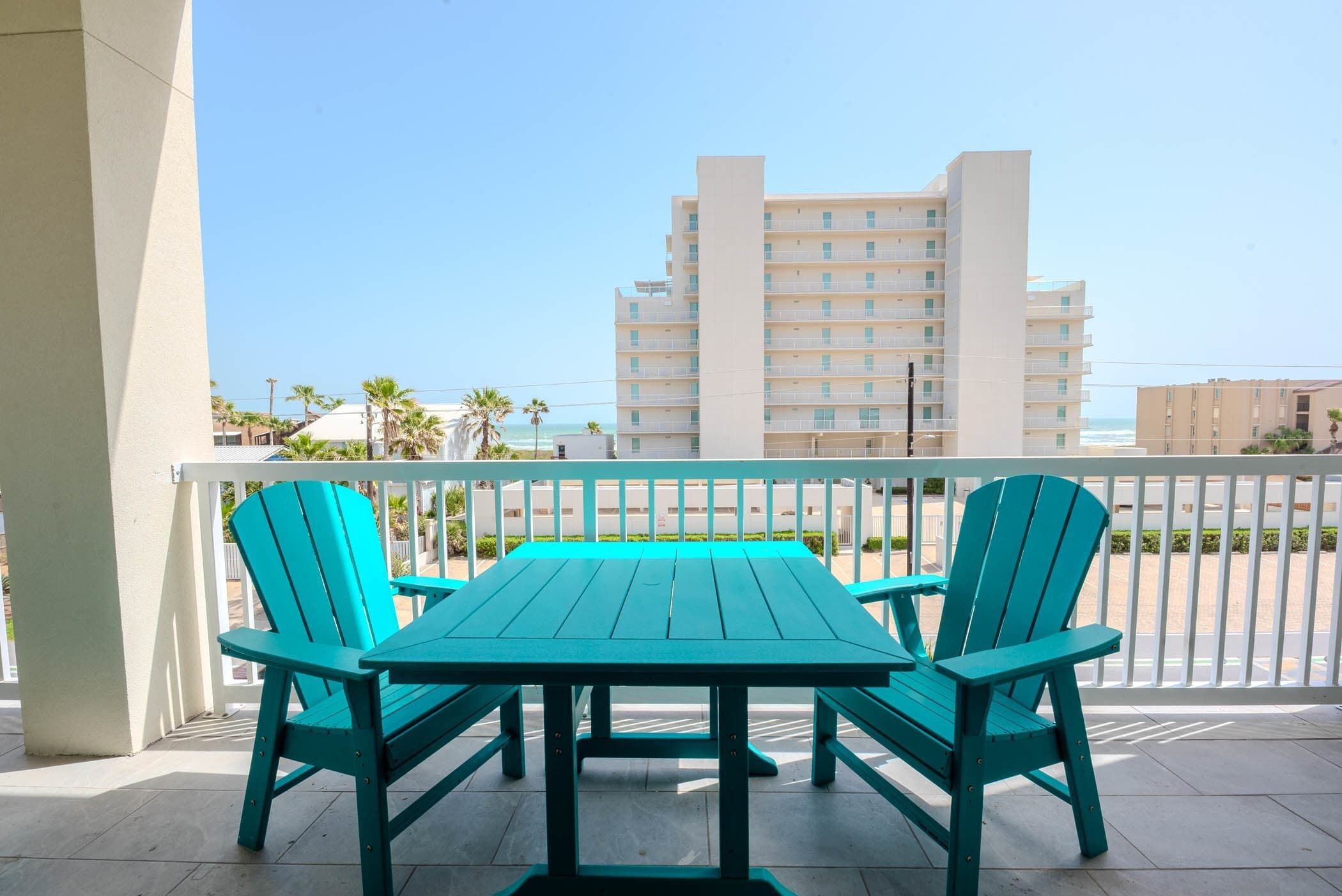 Ein Balkon mit Tisch und Stühlen, der auf ein Gebäude und einen Strand blickt.