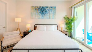 Una habitación de hotel moderna con una cama grande, una mesita de noche con lámparas, una silla, un ventanal amplio y un cuadro en la pared.