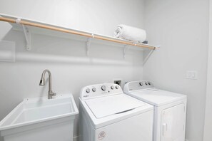 A laundry room with a sink, two white Whirlpool washers, and a wooden shelf with a rolled towel.