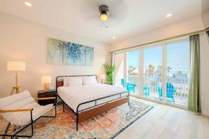 A bedroom with a bed, a chair, a nightstand, a lamp, a painting, and a balcony with chairs.