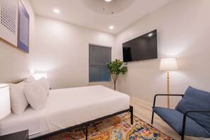 A modern bedroom with a bed, a chair, a TV, and a window with blinds.