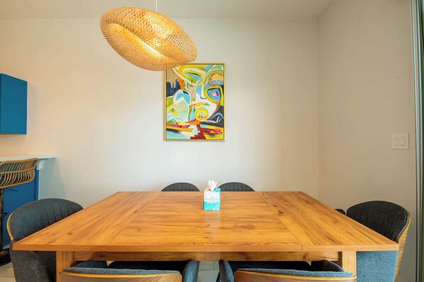 A wooden dining table with four chairs, a colorful abstract painting above it, and a woven light fixture.