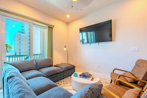 A modern living room with a grey sectional sofa, a round coffee table, and a wooden armchair. A flat-screen TV is mounted on the wall.