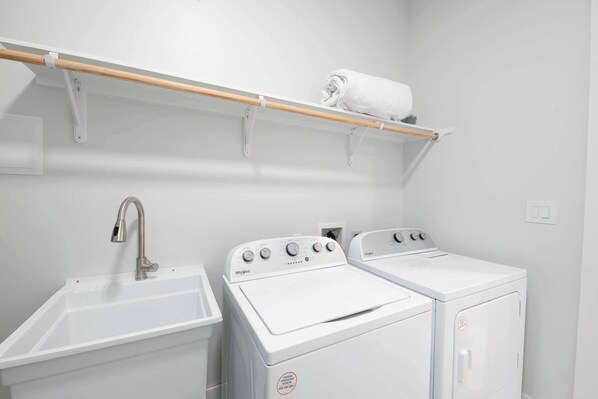 A laundry room with a sink, two white Whirlpool washers, and a wooden shelf with a rolled towel.